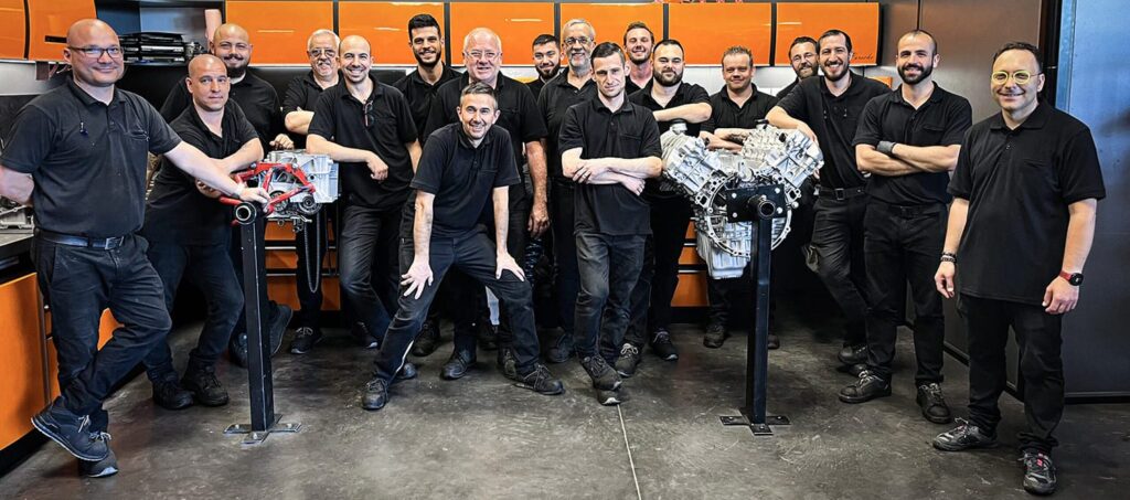 Les techniciens du service production de FLAT autour de moteurs flat-six sur supports, dans l'atelier moteur aux établis orange