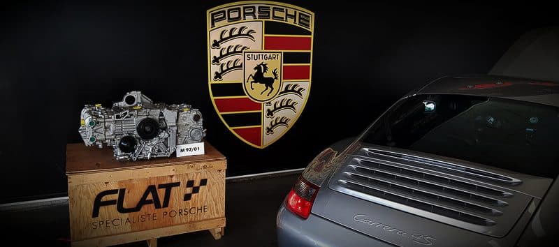 Moteur flat-six reconditionné sur sa caisse FLAT à côté d'une Porsche 911 type 997 grise, devant l'écusson Porsche