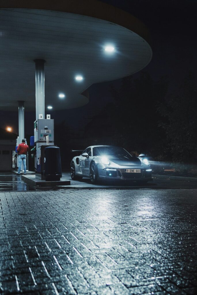 Porsche 911 blanche sous l'auvent éclairé d'une station-service, sol mouillé, de nuit