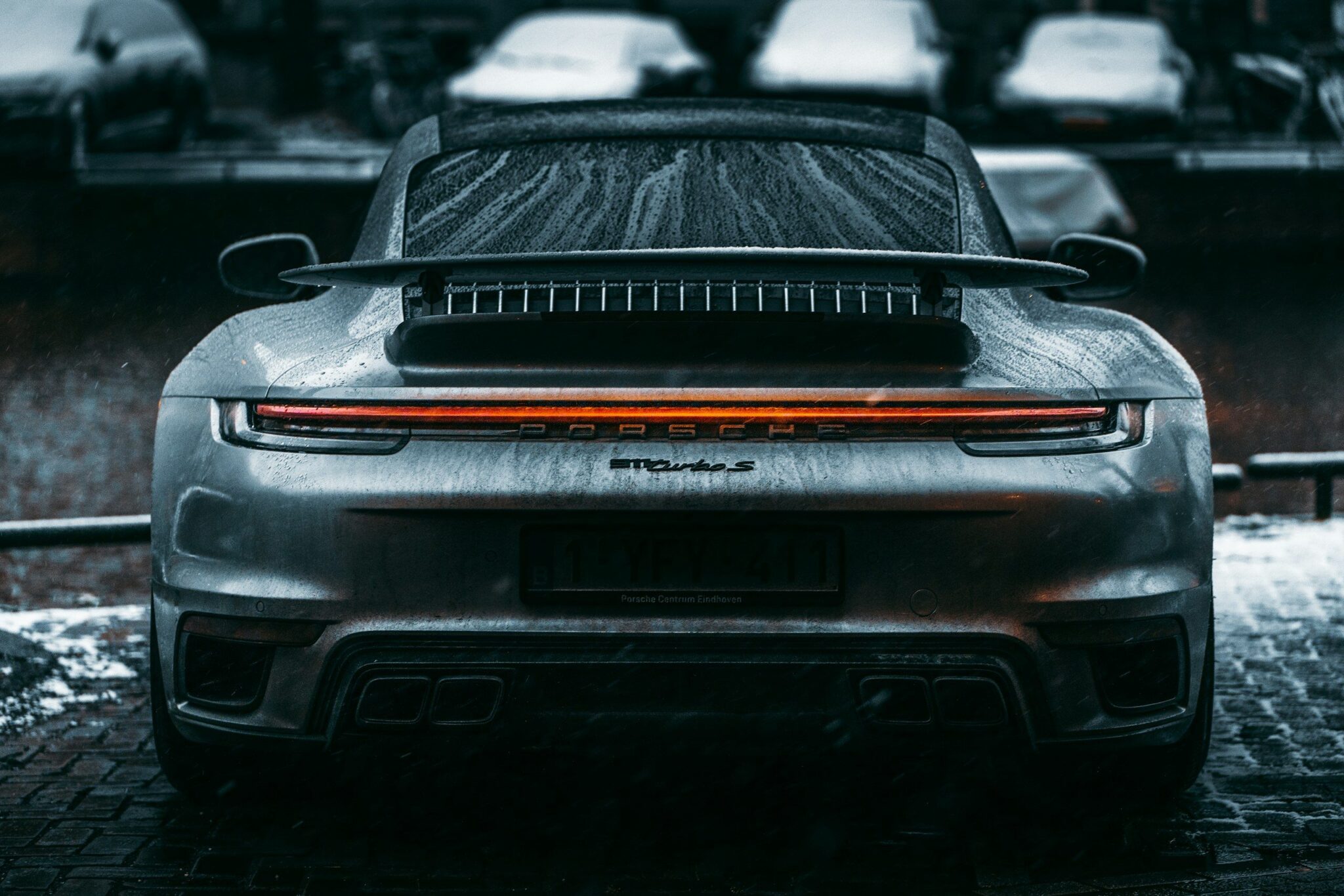 Arrière d'une Porsche 911 Turbo type 992 grise couverte de gouttes de pluie, bandeau lumineux allumé