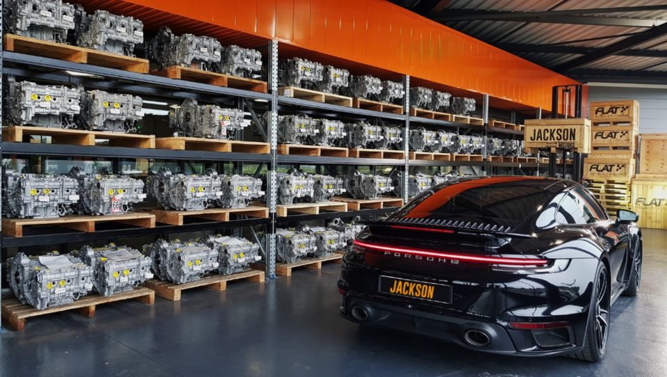 Rayonnages de moteurs flat-six reconditionnés sur palettes et Porsche 911 Turbo noire dans l'entrepôt FLAT
